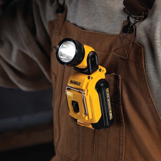 A DEWALT DCL510 portable LED work light clipped onto the front pocket of brown overalls. The light is turned on and provides hands-free illumination.