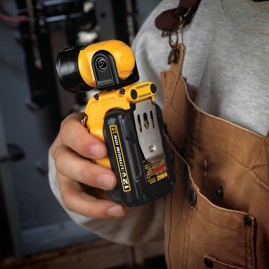 A person wearing a brown work apron and grey shirt is holding a DEWALT DCL510 12V Lithium Ion cordless LED work light in their hand. The tool has a compact, yellow and black design, and is clipped to the person's pocket.
