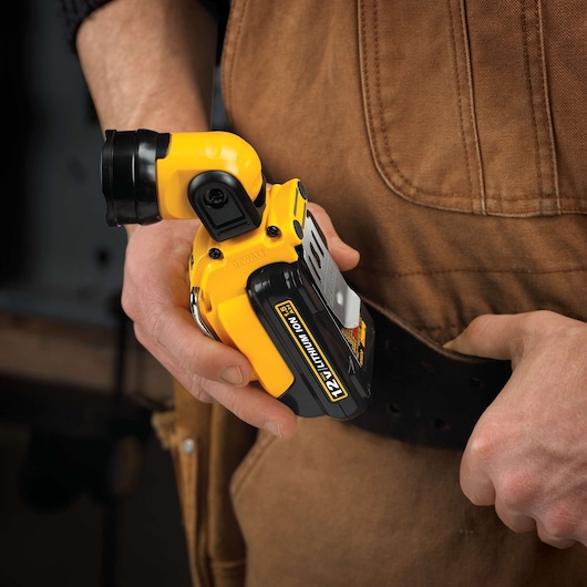 A person wearing brown work overalls holds a yellow DEWALT DCL510 12V lithium-ion cordless LED spotlight, attaching it to a black tool belt.