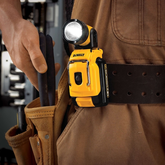 A DEWALT DCL510 portable LED work light clipped onto the belt of a person wearing a brown work apron. The person is holding tools in a pouch beside the light.