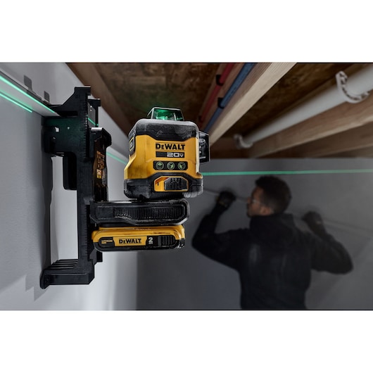 A DEWALT 20V laser level tool mounted on a wall, projecting a green horizontal laser line. A person is in the background working on the wall, with their face blurred.