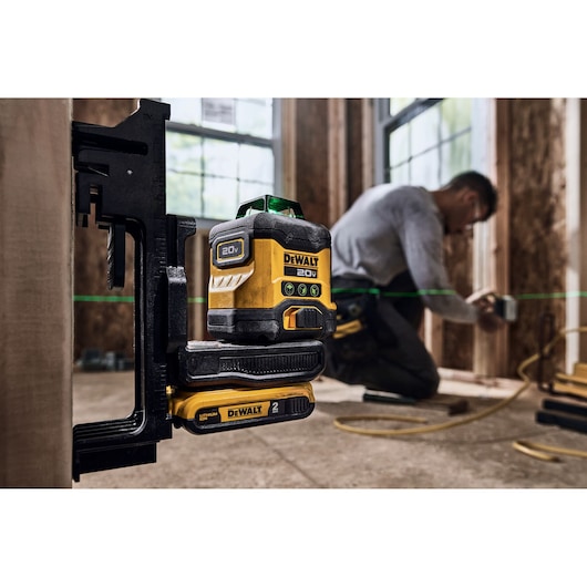 A DEWALT 20V laser level tool mounted on a clamp, projecting green laser lines across a construction site. A person is working in the background on wooden framing.