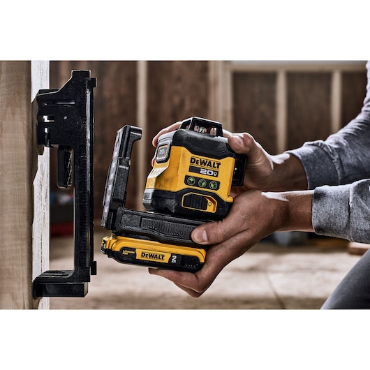 A person is holding a yellow and black DEWALT 20V laser level tool, using it to align with a wall-mounted bracket in a construction setting.
