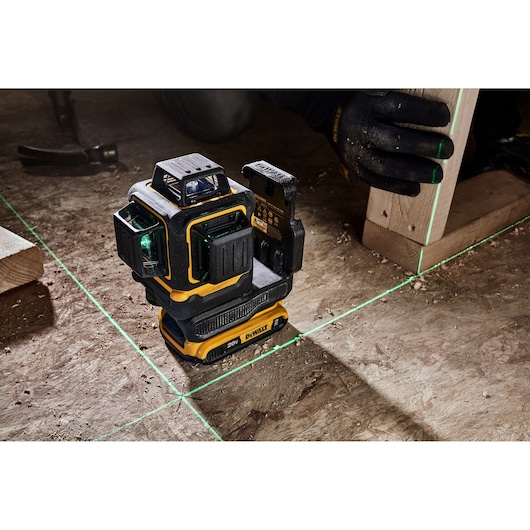 A DEWALT 20V laser level tool projects bright green laser lines onto a wooden floor. A person wearing black gloves is aligning a piece of wood using the laser lines.