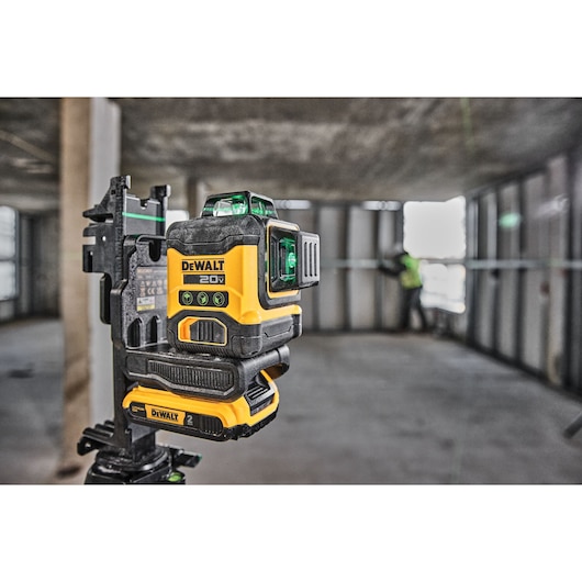 A DEWALT 20V green laser level is mounted on a tripod in a construction site, with a worker in a safety vest visible in the background adjusting metal wall studs.