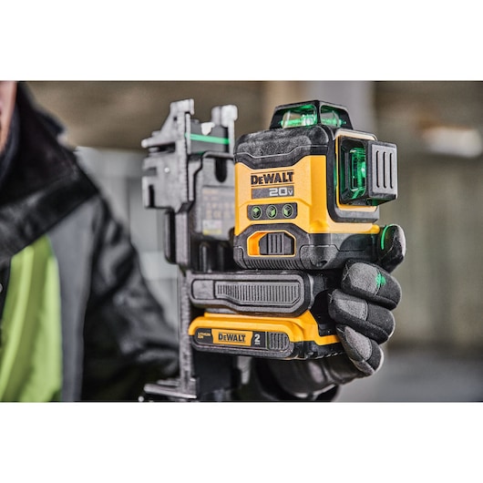 A close-up of a person holding a DEWALT 20V green laser level tool, model DCLE34031B, in a construction setting. The tool is yellow and black with a rechargeable battery attached.