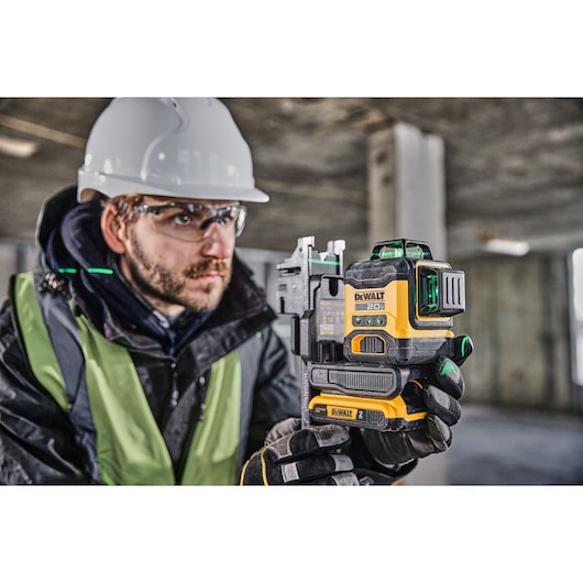A person wearing safety gear holds a DEWALT 20V green laser level device at a construction site. The focus is on the tool, which is used for accurate alignment and leveling.