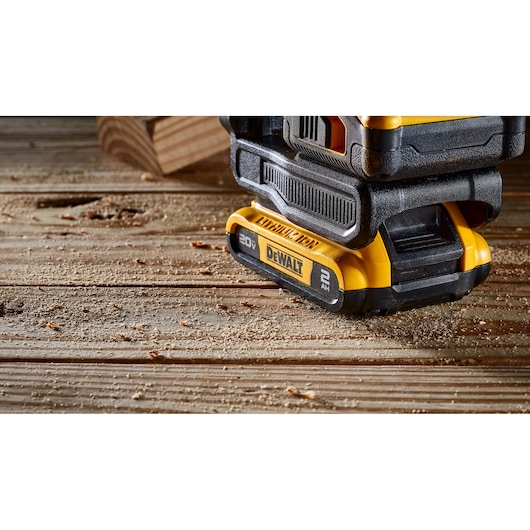Close-up of a DEWALT 20V power tool battery resting on a wooden surface with sawdust, showing part of the tool and a piece of wood in the background.