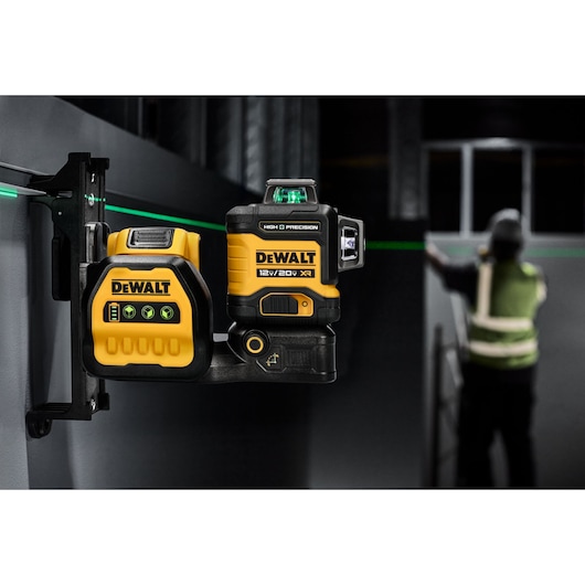 A DEWALT 12V/20V XR high precision green laser level is mounted on a wall bracket. It emits a visible green laser line across the workspace. The device is yellow and black, featuring DEWALT branding. In the background, a person wearing a reflective vest is working near the laser line.
