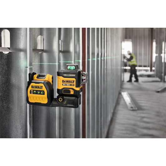 A DEWALT laser level device is mounted on a metal construction wall, projecting a green horizontal laser line across the room. In the background, a person wearing a safety vest is working in the unfinished industrial space.