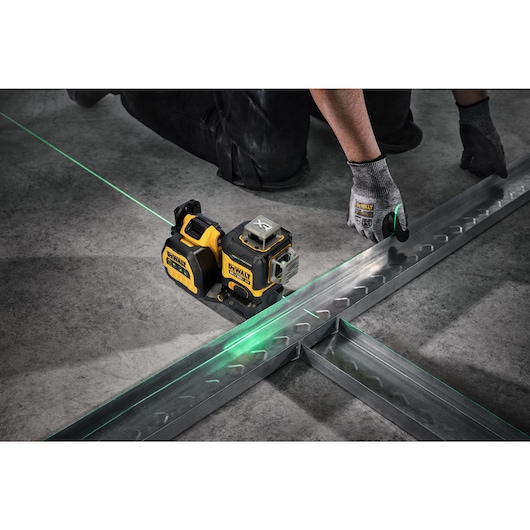 A DeWalt laser level device projects a green laser line onto a concrete floor, assisting a worker wearing gloves to align metal framing pieces.