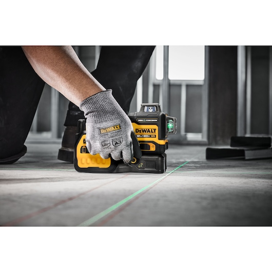A person wearing a grey work glove is setting up a DEWALT 12V/20V XR laser level on a concrete floor, projecting bright green laser lines to assist in alignment. The tool is ideal for precision layout tasks in construction.
