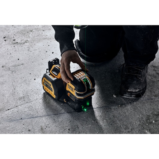 A person using a DEWALT laser level tool on a concrete floor. The tool emits a green laser line for precise leveling and alignment.