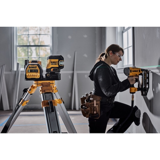 A DEWALT laser level set up on a tripod in a construction room, with a person using a DEWALT power tool to work on a wall. The person’s face is blurred and they are wearing a tool belt.