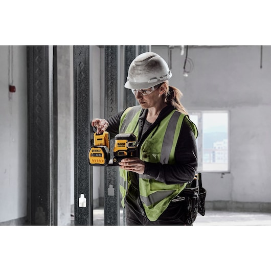 A construction worker wearing a hard hat and reflective vest operates a DEWALT laser level tool at a building site. The tool is yellow and black, and the worker is in an unfinished room with metal studs and construction materials.