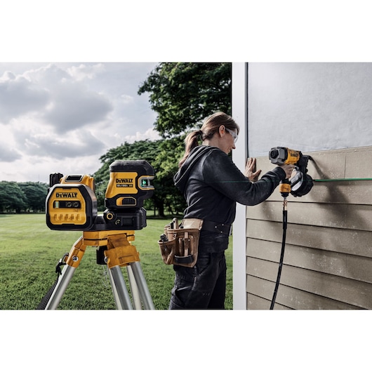 A person using a DEWALT laser level and power tool to install siding on the exterior wall of a building, with the DEWALT laser level mounted on a tripod in the foreground. The person is wearing a tool belt and working outdoors.