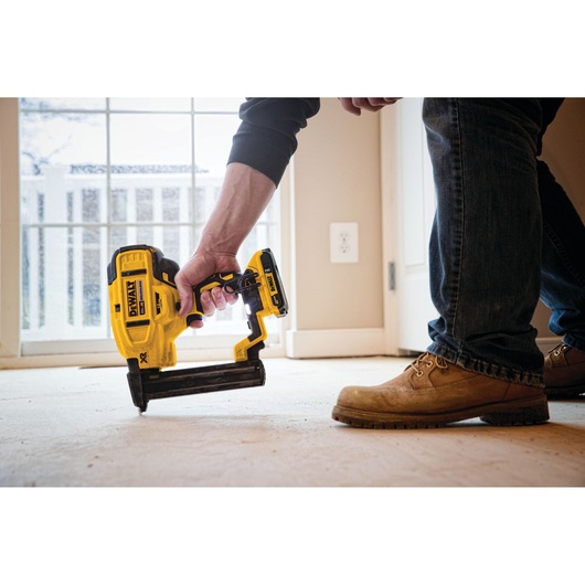 A person using a DEWALT DCN681B cordless nailer on a floor in a well-lit room, wearing work boots and jeans.