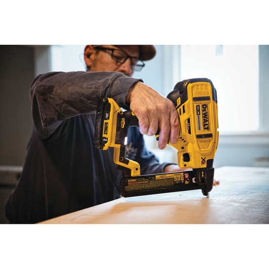 A person using a DEWALT DCN681B cordless nailer tool on a piece of wood indoors. The focus is on the tool and the person's hand.