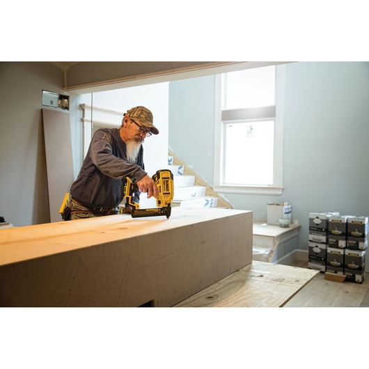 A person using a DEWALT cordless nailer to fasten wood on an indoor construction site. Boxes of supplies are visible in the background, along with a staircase and large window.