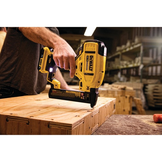A person uses a DEWALT cordless nailer to fasten wooden boards in a workshop. The tool is yellow and black, with visible branding, and is being applied to plywood on a workbench.