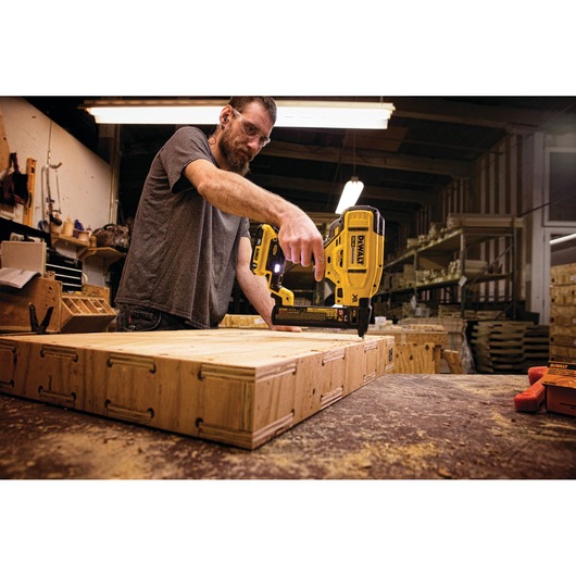 A person using a DEWALT cordless nailer tool to work on a wooden crate in a workshop.