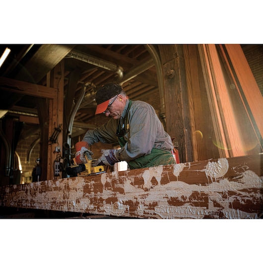 A person wearing work gloves and protective clothing uses a DEWALT cordless hand planer to smooth a large wooden beam inside a workshop. The beam has visible patches of paint or finish throughout its surface.
