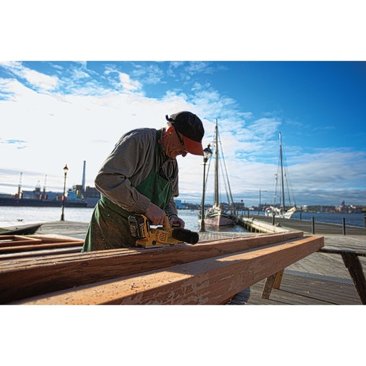 A person wearing a cap and apron planes a wooden plank with a DEWALT cordless hand planer outdoors near a waterfront with sailboats and industrial buildings in the background.