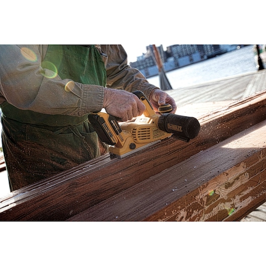 A person using a DEWALT cordless planer to smooth the surface of a wooden beam outdoors near water.