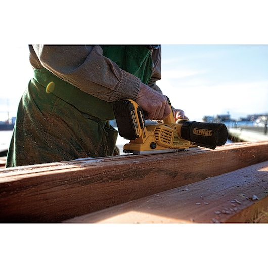 A person using a DEWALT cordless planer to smooth a piece of wood outdoors.