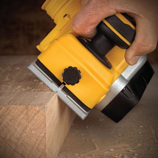 A close-up of a hand using a yellow cordless planer tool to smooth the edge of a wooden block, with wood shavings visible on the surface.