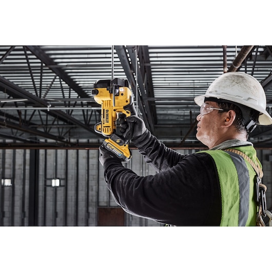 A construction worker wearing a helmet and safety vest uses a DEWALT DCS350B tool to cut a metal rod in an industrial setting.