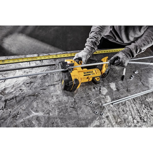 A person wearing gloves uses a yellow DEWALT cordless threaded rod cutter to cut metal rods on a workbench. Several rods, nuts, and a measuring tape are visible on the table.