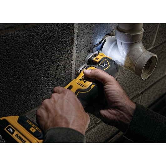 Close-up of a person using a DEWALT cordless oscillating multi-tool to cut or sand around a plastic plumbing pipe mounted on a concrete wall.