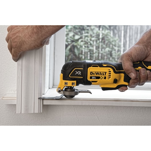 A person using a DEWALT 20V XR oscillating multi-tool to trim a piece of white wood molding near a window.