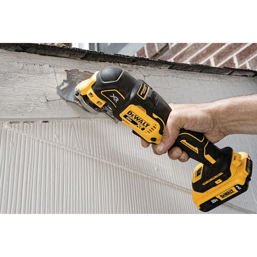 A DEWALT XR cordless oscillating multi-tool is being used to cut or sand a painted wooden surface near the roofline of a building.