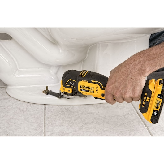 A person uses a DEWALT XR 20V cordless oscillating multi-tool to cut a metal bolt at the base of a white toilet on a tiled floor.