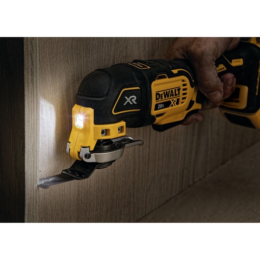 A close-up of a DEWALT 20V XR cordless oscillating multi-tool being used to cut into a wooden surface. The tool is handheld and has an LED light illuminating the work area.