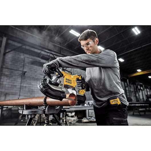 A person using a DEWALT DCS374B portable band saw to cut a metal pipe in an industrial workshop. The person's face is blurred for privacy.