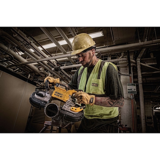 A worker wearing a yellow safety helmet, gloves, and a high-visibility vest is using a DEWALT DCS374P2 cordless band saw in an industrial setting with exposed pipes and machinery in the background.