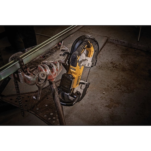 A DEWALT cordless band saw is shown cutting a metal pipe held in a clamp in a workshop setting.