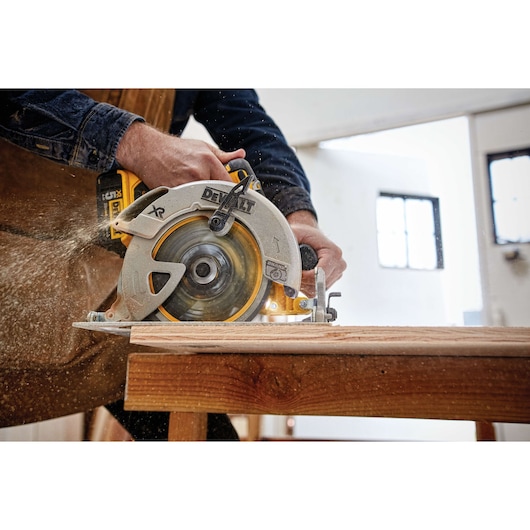 A person using a DEWALT cordless circular saw to cut a piece of wood in a workshop. Sawdust is visible as the tool operates.