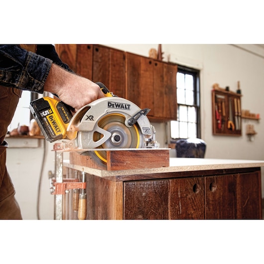 A person using a DEWALT cordless circular saw to cut a piece of wood on a workbench in a woodworking shop.