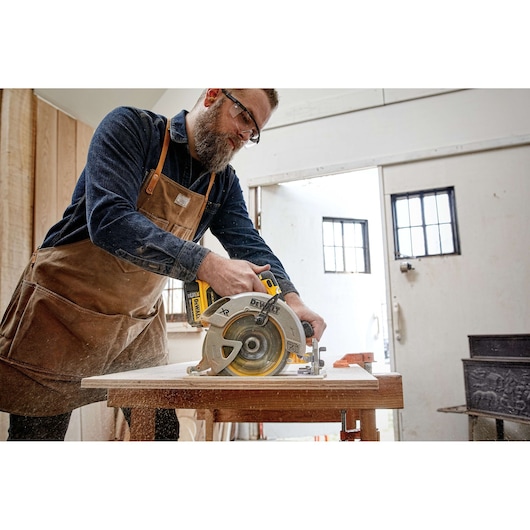 A person wearing a work apron and dark shirt is using a DEWALT cordless circular saw to cut a piece of wood in a workshop. The saw is yellow and silver, and sawdust is visible from the cutting process.