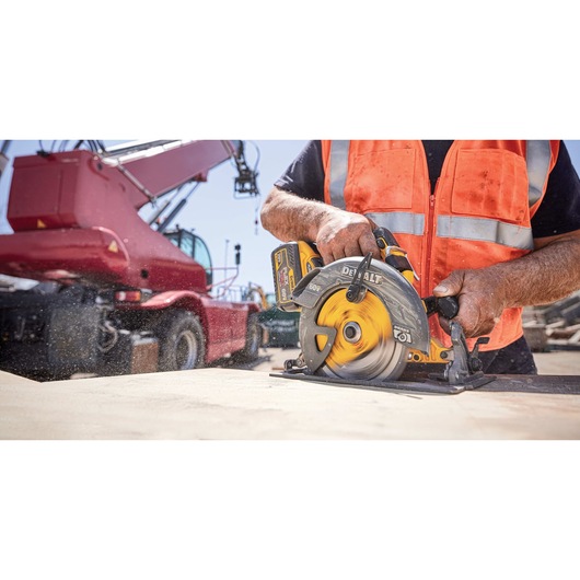 A person wearing an orange safety vest is using a yellow DEWALT circular saw to cut wood on a construction site, with a large red construction vehicle in the background.