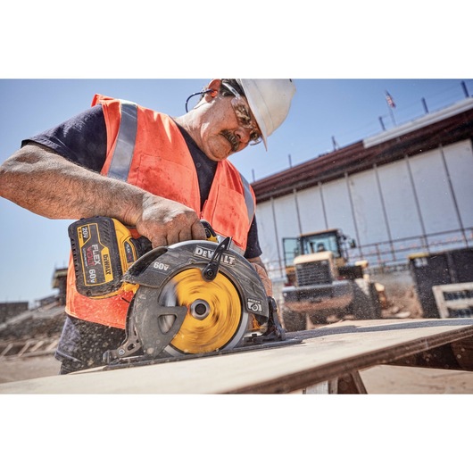 A person wearing an orange safety vest and helmet is using a DEWALT DCS575B 60V cordless circular saw to cut wood at a construction site.