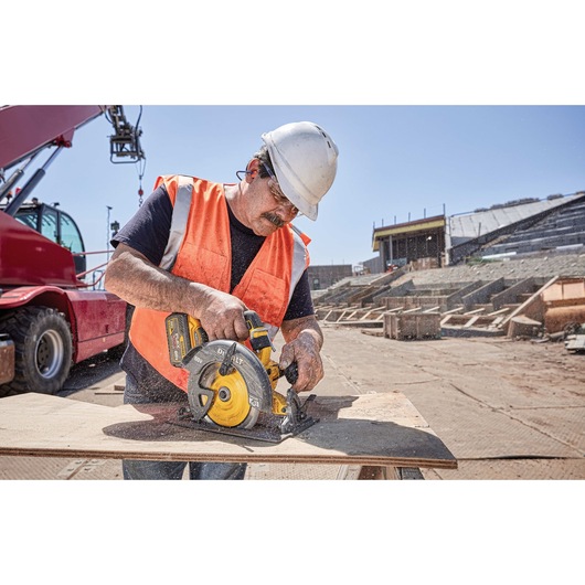 A construction worker wearing a white hard hat and orange safety vest uses a DEWALT circular saw to cut a wooden board at a construction site. There is construction equipment and a partially built structure in the background.