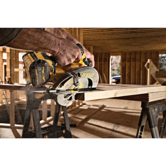 A close-up of a person using a DEWALT cordless circular saw to cut a piece of wood inside a building under construction.