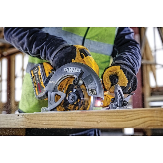 A close-up of a person using a DEWALT circular saw to cut a piece of wood. The person is wearing yellow work gloves and a bright safety vest, with a construction site visible in the background.