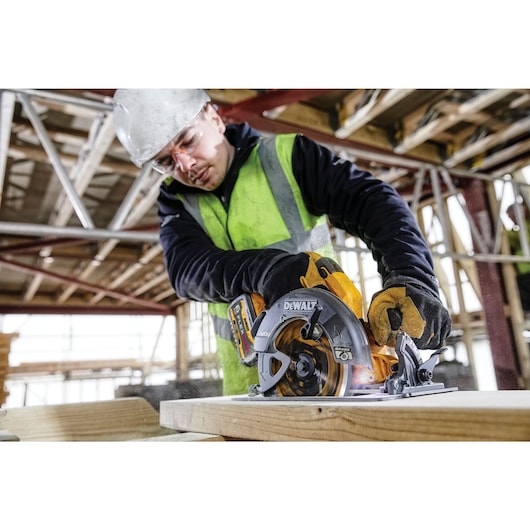 A person wearing a safety vest and gloves is using a DeWalt circular saw to cut wood at a construction site. Their face is blurred for privacy, and scaffolding is visible in the background.
