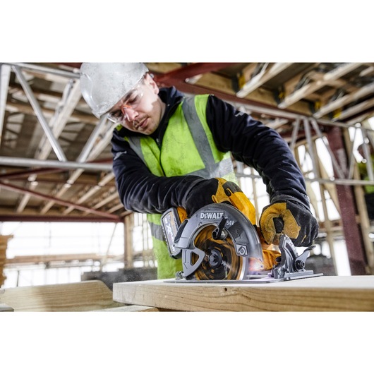 A close-up image of a person wearing gloves and a reflective vest operating a DEWALT DCS578T2 cordless circular saw to cut a wooden plank. The saw features a 60V Flexvolt battery and spinning blade in a construction environment.